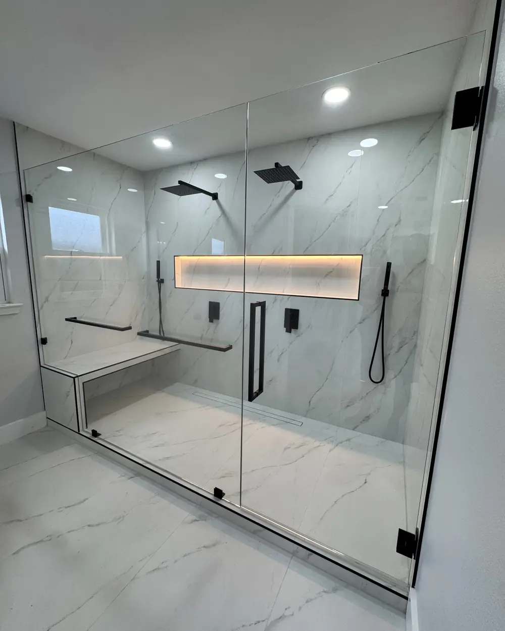 Walk-in shower with large marble-look tile, a lit wall niche, built-in bench and black fixtures
